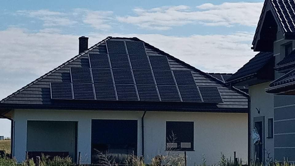 Ciemne panele na dachu domu z szarą dachówką, widok na elewację i błękitne niebo z chmurami.