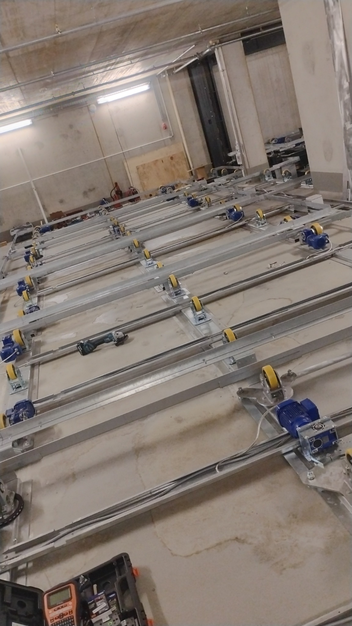 Rozległa konstrukcja sufitowa z szynami, wózkami i silnikami elektrycznymi, widoczna w surowym, przemysłowym wnętrzu z narzędziami na podłodze.