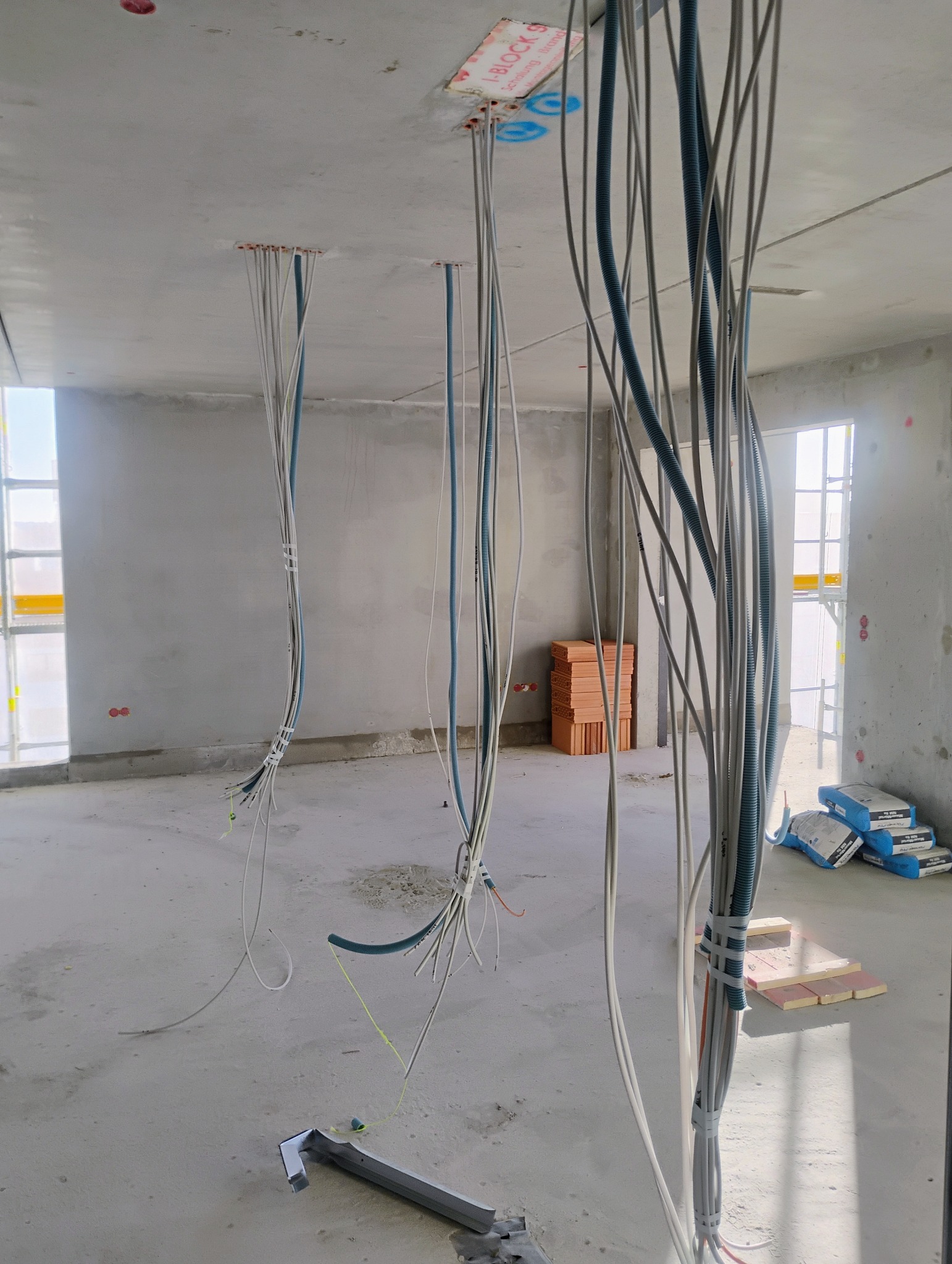 Wiązki przewodów elektrycznych zwisające z sufitu w surowym stanie deweloperskim, widoczne puszki instalacyjne, cegły i worki z zaprawą w tle, narzędzie na posadzce.
