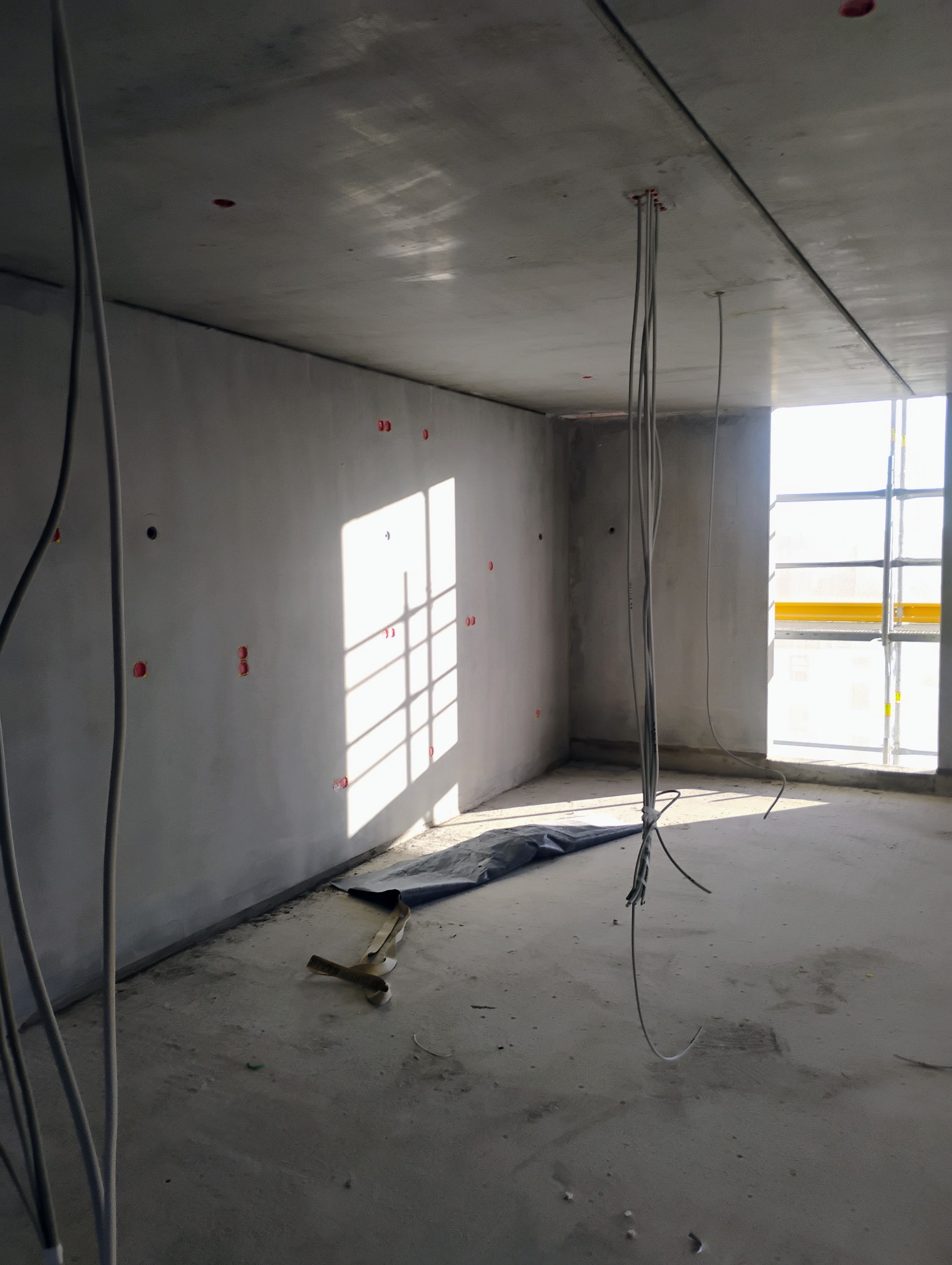 Surowy stan wnętrza z wystającymi przewodami elektrycznymi z sufitu, przygotowane otwory w ścianach, podłoga zabezpieczona folią, widoczne ślady prac budowlanych, wpadające światło słoneczne przez...