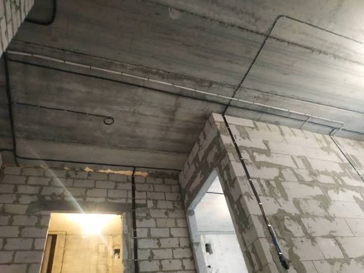 Surowy betonowy strop z widocznymi czarnymi i białymi przewodami elektrycznymi, instalacja w trakcie montażu w budynku w stanie deweloperskim z surowymi ścianami z betonu komórkowego.