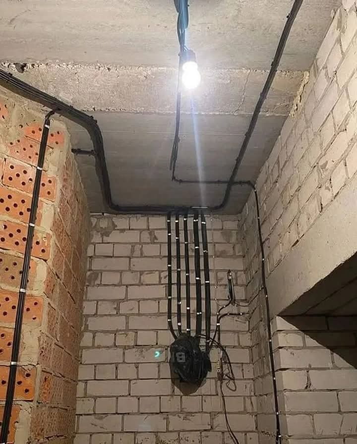 Rozprowadzenie przewodów elektrycznych w peszlach, mocowanych do ceglanej ściany w surowym stanie, widoczne kable pod sufitem i podłączenie do puszki elektrycznej.