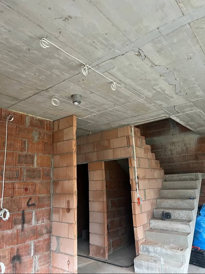 Surowy stan budynku w trakcie prac: widoczne przewody elektryczne poprowadzone po betonowym suficie, ściany z czerwonej cegły, surowe schody betonowe prowadzące na wyższą kondygnację.