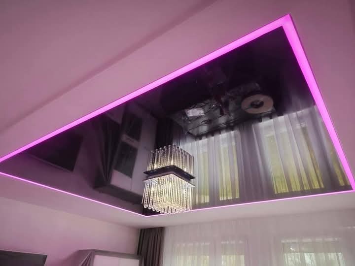 Błyszczący, czarny sufit napinany z obwódką z różowego oświetlenia LED, odbijający wiszący kryształowy żyrandol i okno z firankami.
