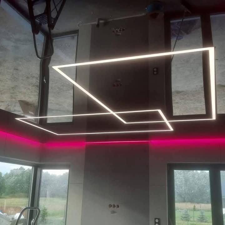 Nowoczesne oświetlenie sufitowe LED w kształcie geometrycznym, z dodatkowym podświetleniem w kolorze różowym, widoczne na tle okien i szarej ściany.
