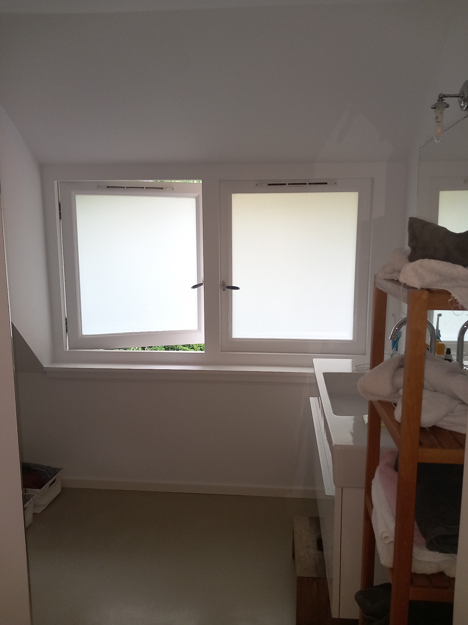 Wnętrze łazienki z oknami z mleczną szybą, drewnianym regałem na ręczniki i białą umywalką. Jedno okno uchylone, widoczna zieleń na zewnątrz. Ujęcie z perspektywy wejścia do łazienki.