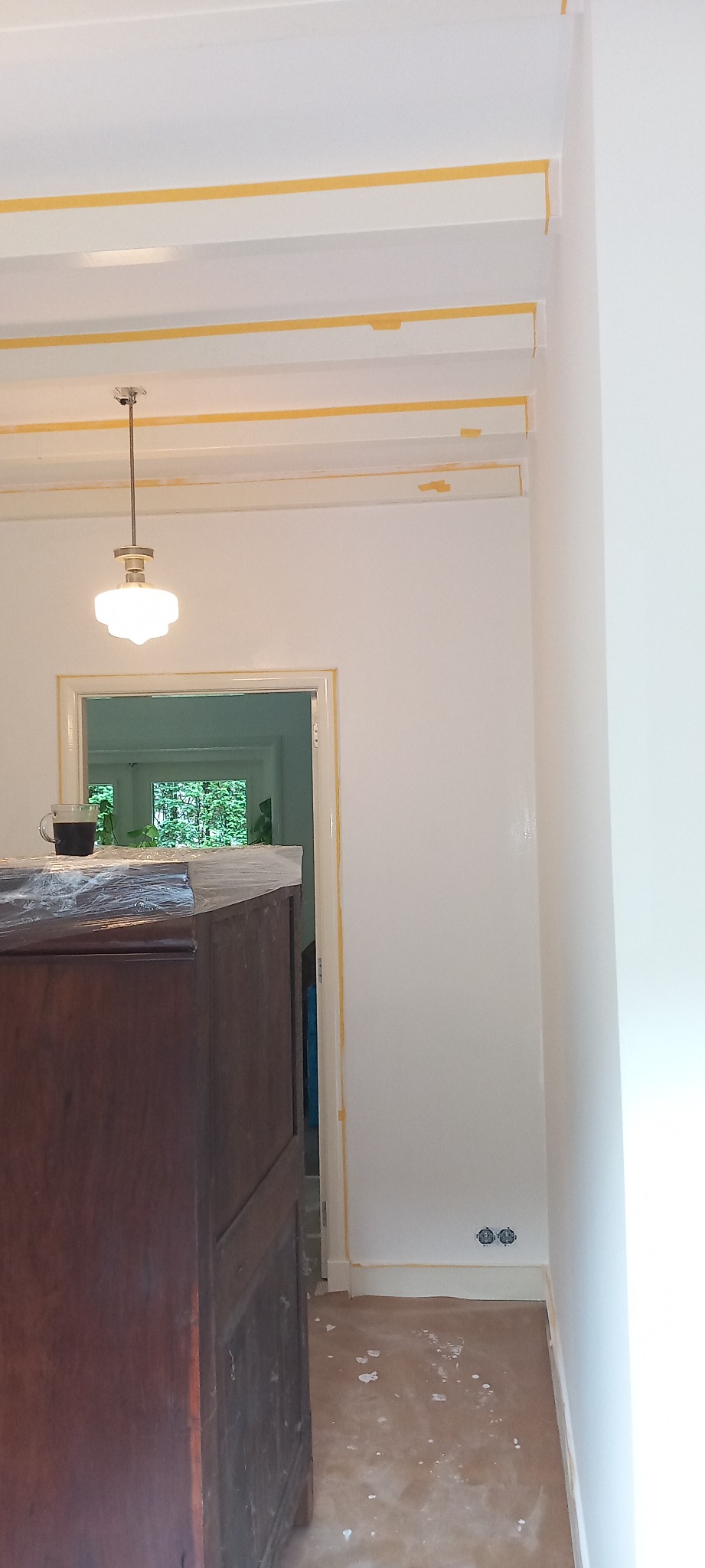 Wnętrze w trakcie malowania: białe ściany z żółtą taśmą malarską na suficie i wokół drzwi, zakryty mebel z filiżanką na wierzchu, widoczne gniazdka elektryczne, ślady farby na podłodze.