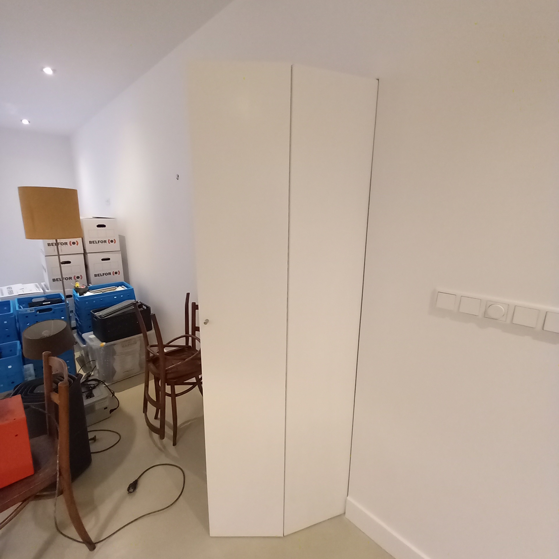 Nowa, biała szafa wnękowa z przesuwnymi drzwiami w trakcie montażu, z widocznymi kartonami i krzesłami w tle, sugerującymi prace wykończeniowe w pomieszczeniu.