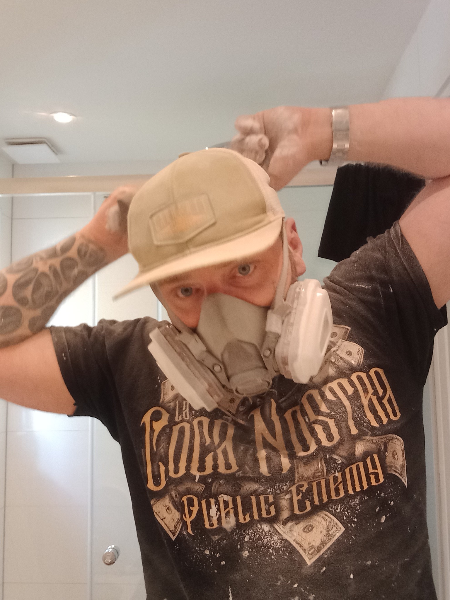 Mężczyzna w czapce i masce ochronnej, ubrudzony pyłem, z tatuażem na ramieniu, poprawia maskę na tle białej łazienki.