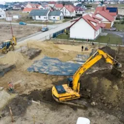 Żółta koparka JCB podczas prac ziemnych na placu budowy, widoczne wykopane fundamenty, w tle zabudowa jednorodzinna i maszyny budowlane.