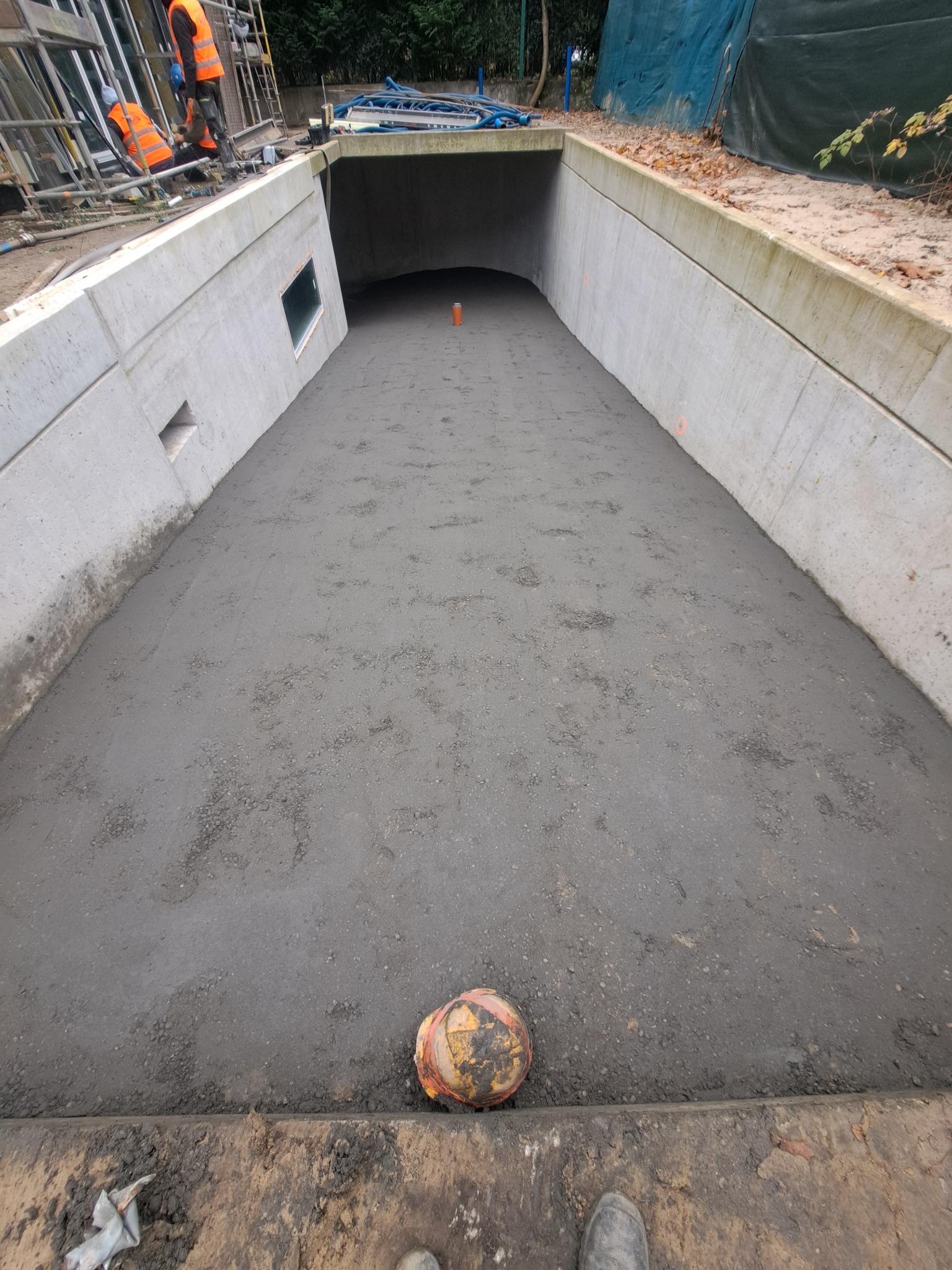 Wnętrze betonowego tunelu w trakcie budowy, świeżo wylana posadzka, widoczne ślady pracy i pomarańczowe znaczniki. W tle robotnicy i rusztowania.