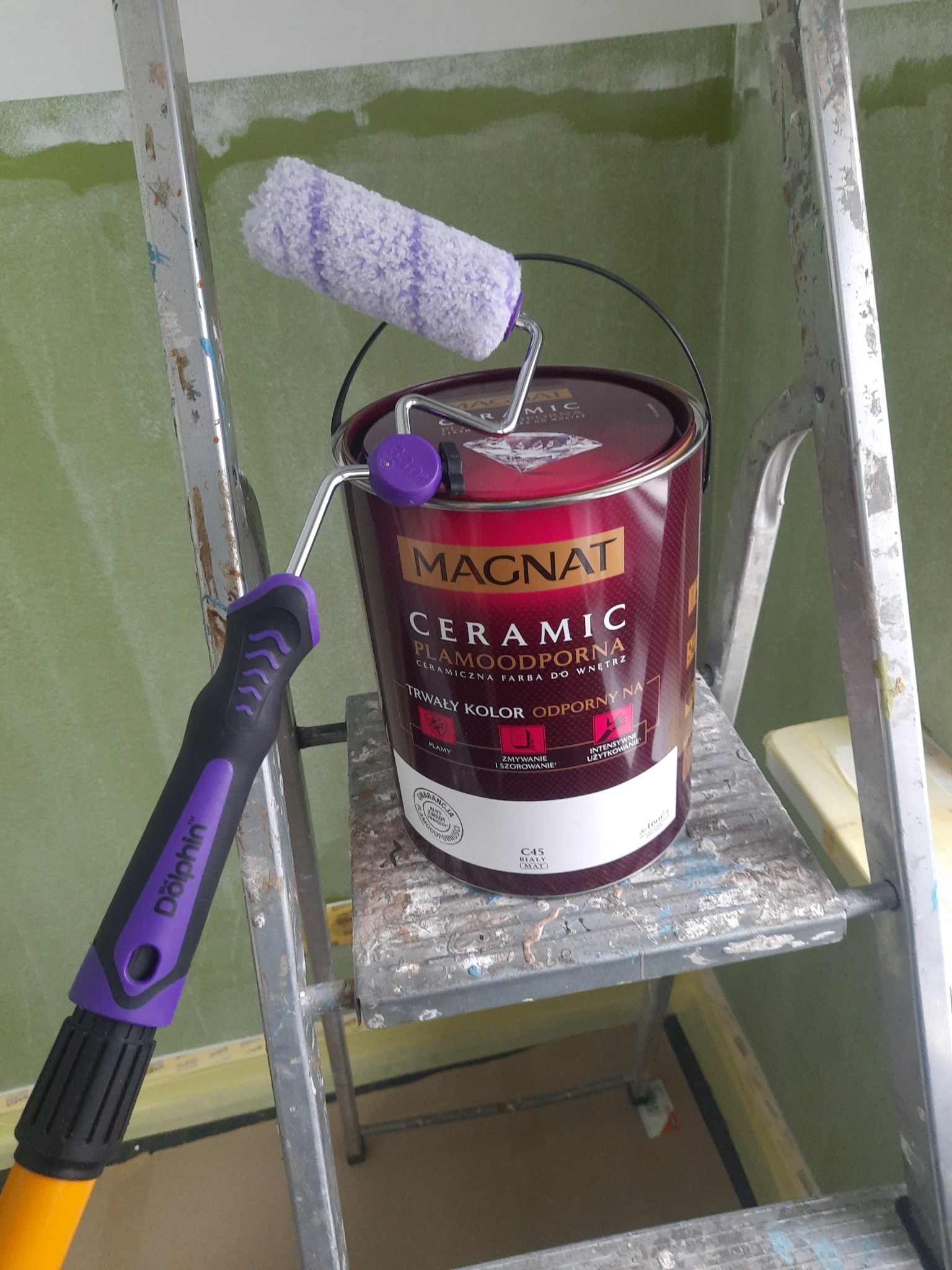 Puszka bordowej farby Magnat Ceramic z fioletowym wałkiem Dolphin na drabinie, ściana w tle pomalowana na zielono.