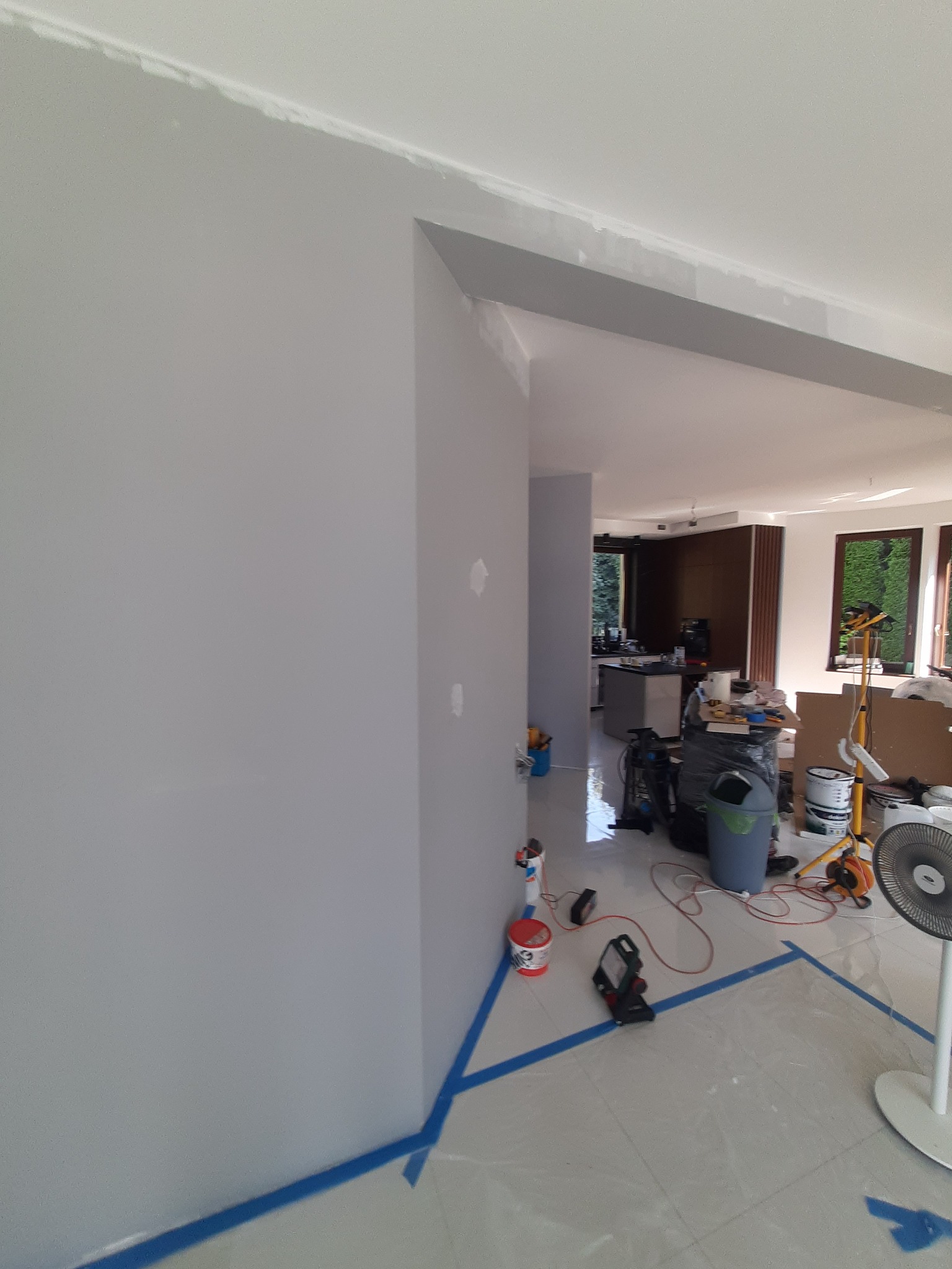 Świeżo pomalowane szare ściany w jasnym wnętrzu domu w trakcie prac wykończeniowych, z widocznymi narzędziami malarskimi, taśmą malarską na podłodze i niedoskonałościami w górnym rogu ściany.