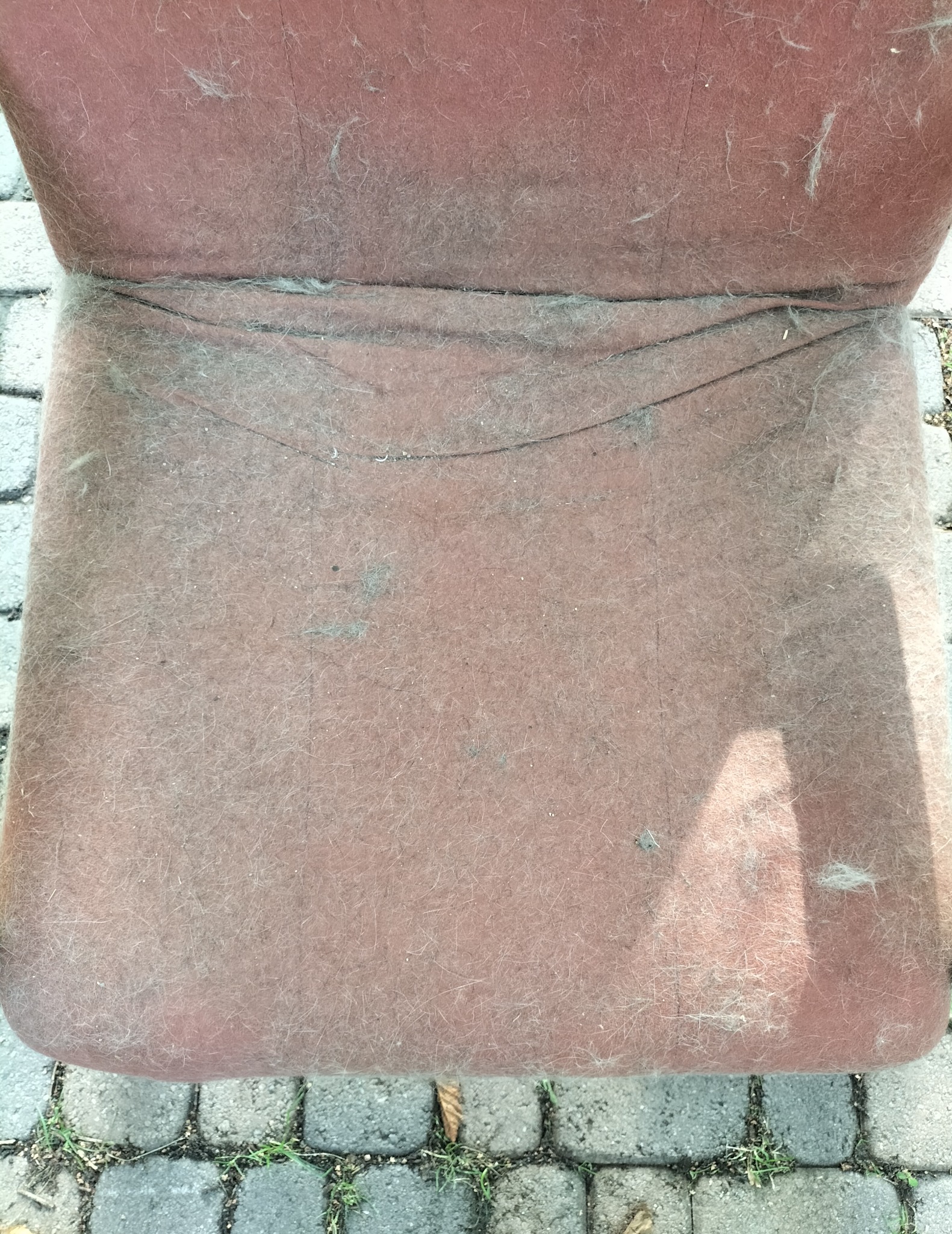 Widok z góry na mocno zabrudzone, brudno-różowe krzesło tapicerowane z widocznymi kłębkami kurzu i sierści, stojące na szarej kostce brukowej.