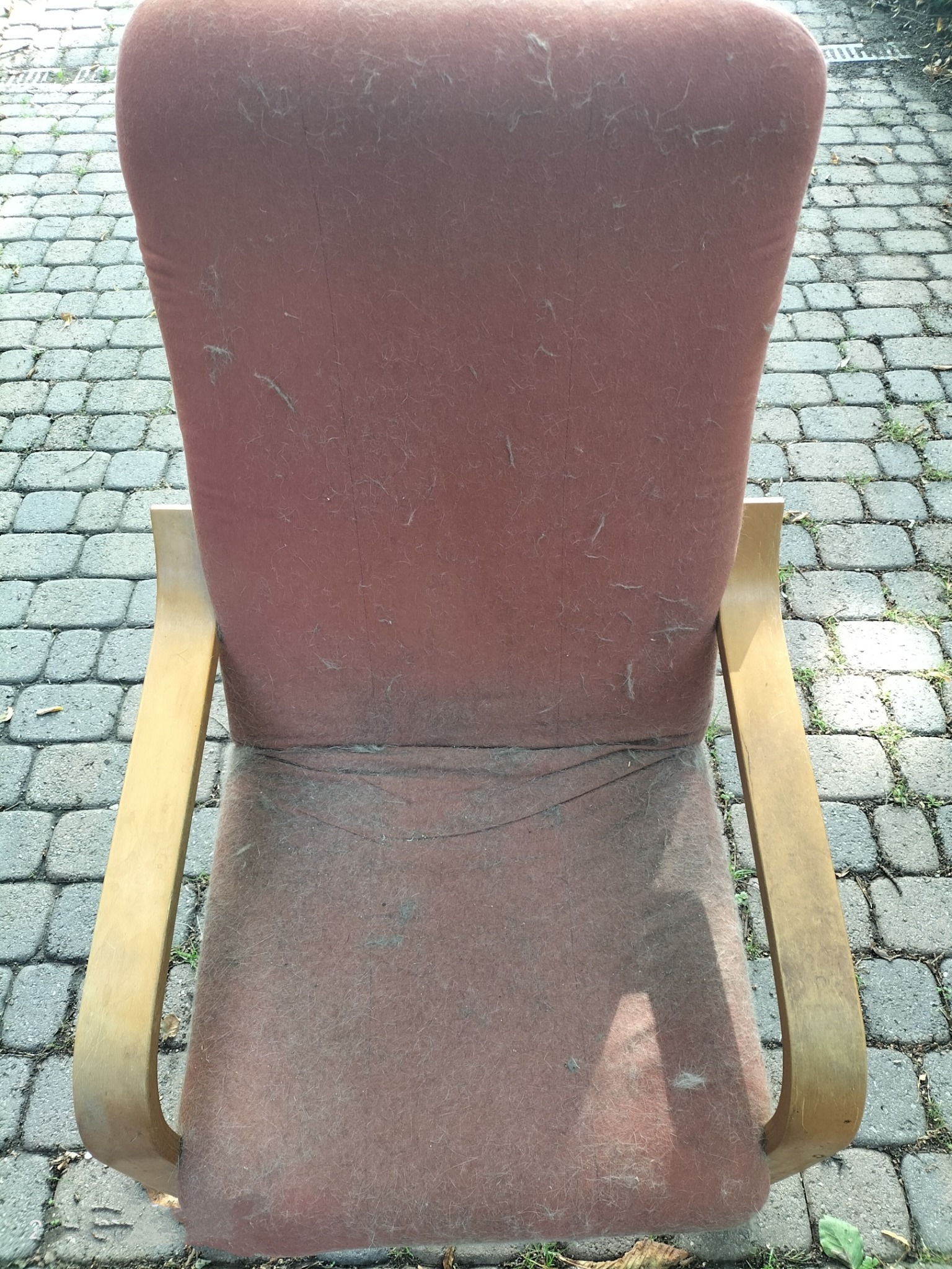 Widok z tyłu na mocno zakurzony, czerwono-brązowy fotel tapicerowany z drewnianymi podłokietnikami stojący na brukowanej powierzchni.