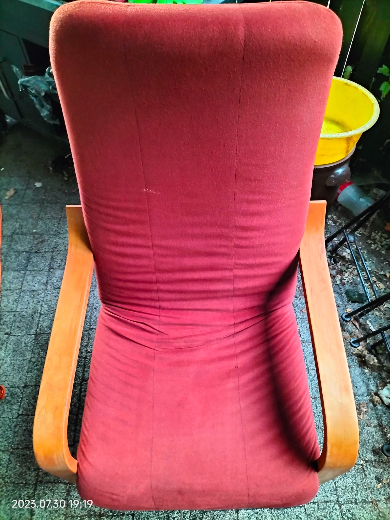 Czerwony fotel tapicerowany z drewnianymi podłokietnikami, widok z tyłu, widoczne ślady użytkowania i żółte wiadro w tle.