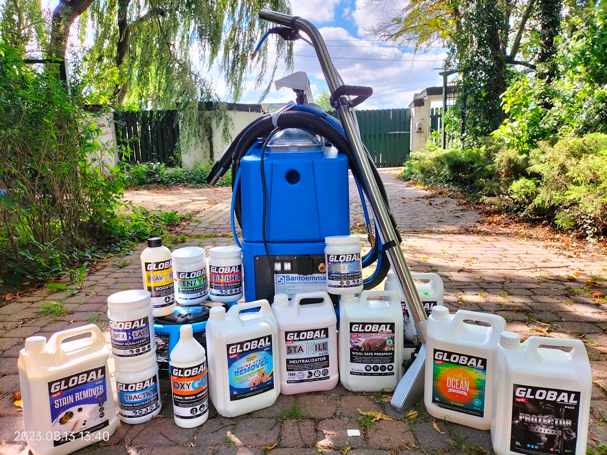 Profesjonalny sprzęt do czyszczenia tapicerki i dywanów marki Santoemma, otoczony różnorodnymi środkami czyszczącymi marki Global, ustawiony na brukowanej ścieżce w ogrodzie.