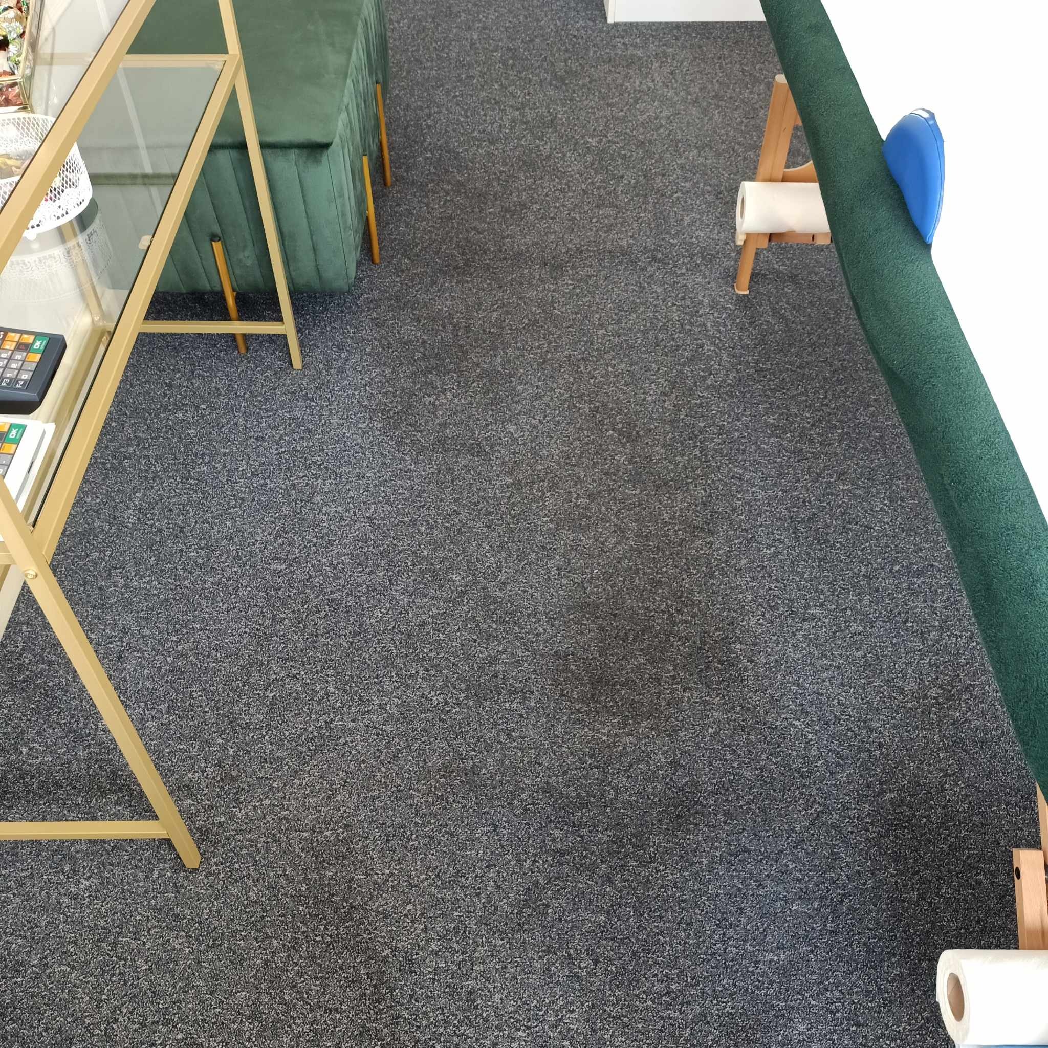 Szary dywan z krótkim włosiem widziany z góry, fragment regału z metalową ramą w kolorze złotym, fragment zielonej sofy i rolka papieru na drewnianym stojaku.