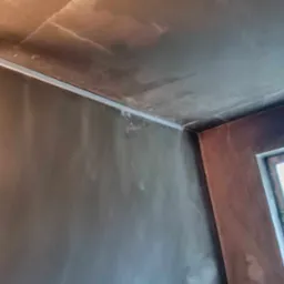 Szare ściany i sufit w trakcie wykańczania, widoczna listwa przypodłogowa i okno z brązową ramą.