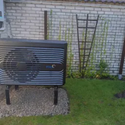 Czarna jednostka zewnętrzna z logo FOX na szarym żwirze, zamontowana przy jasnej ceglanej ścianie, z metalową siatką i pnączami w tle.