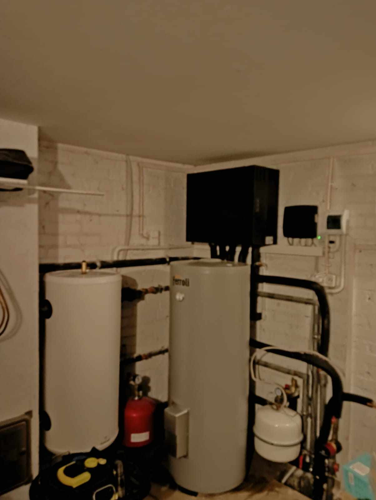 Dwa zbiorniki buforowe, jeden marki Ferroli, z czarną skrzynką i przewodami, zainstalowane w piwnicy z pomalowanymi ceglanymi ścianami.