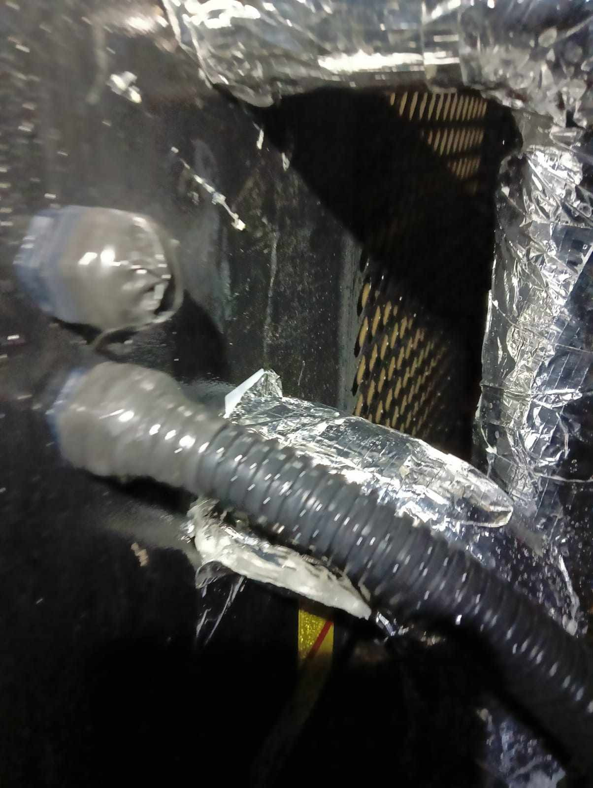 Szczegółowe ujęcie uszczelnionego przewodu wentylacyjnego z folią aluminiową i taśmą izolacyjną, widoczne elementy instalacji w ciemnym otoczeniu.