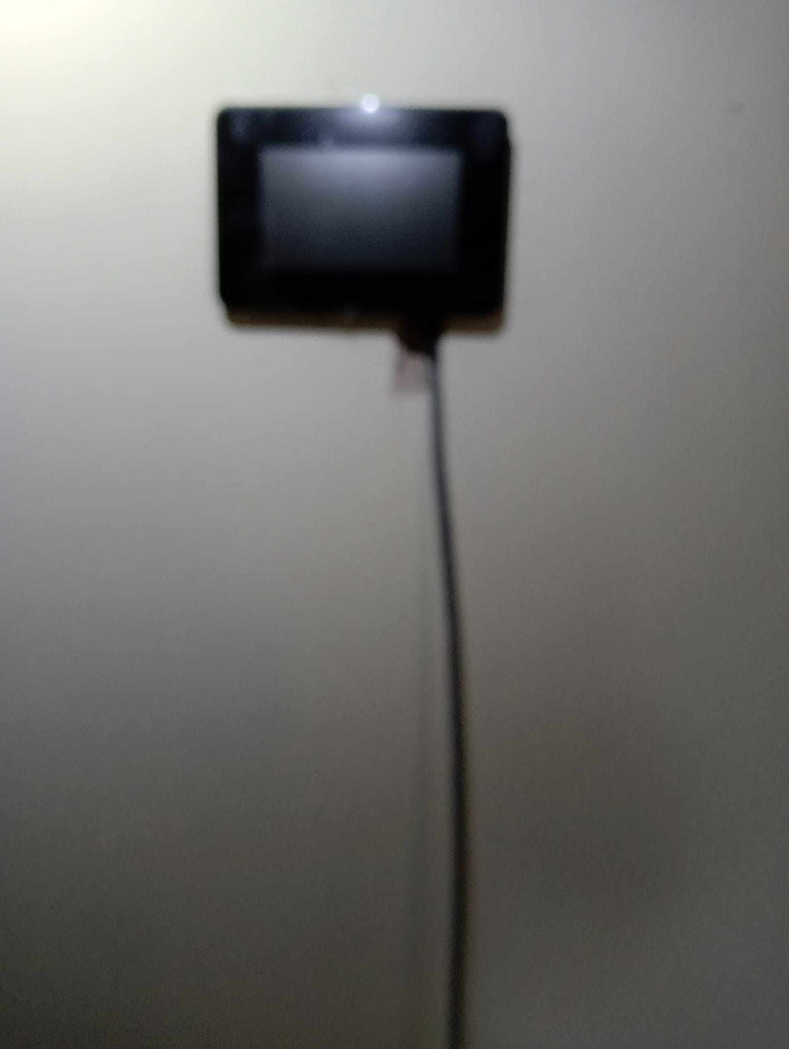 Czarny panel sterowania, prawdopodobnie do pompy ciepła, zamontowany na jasnej ścianie, z widocznym przewodem. Rozmazane zdjęcie.