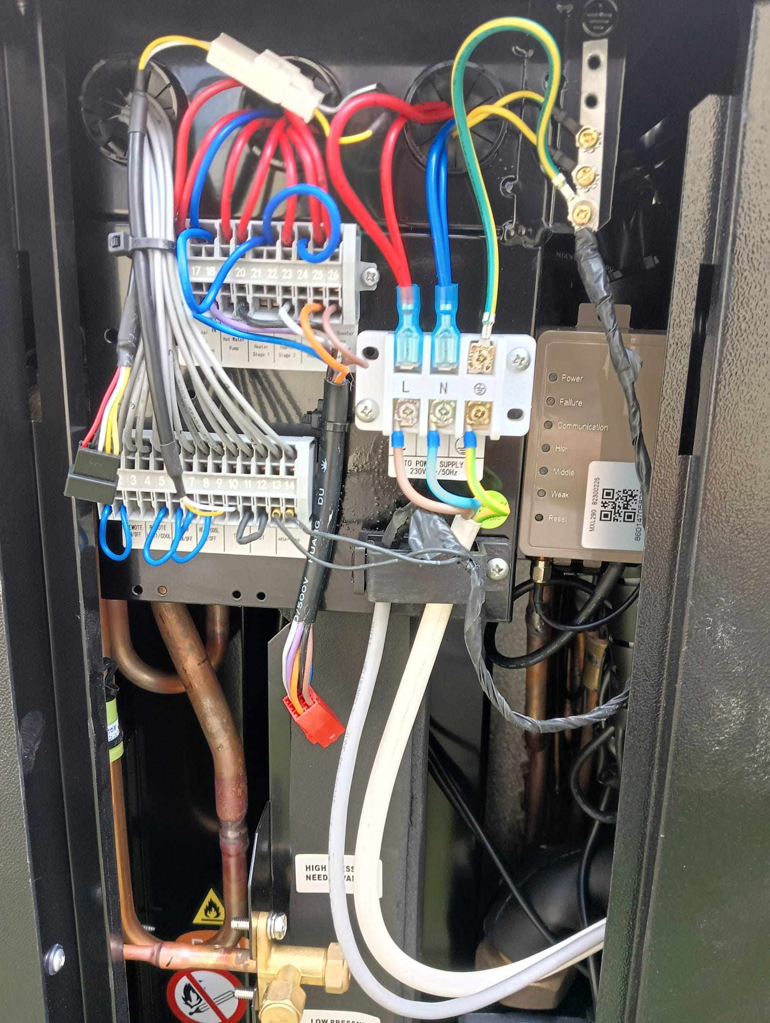 Wnętrze urządzenia z widocznymi elementami elektrycznymi: złączki, kable w różnych kolorach, moduł sterujący z diodami LED i kodem QR oraz miedziane rurki. Widoczne ostrzeżenia o wysokiej...