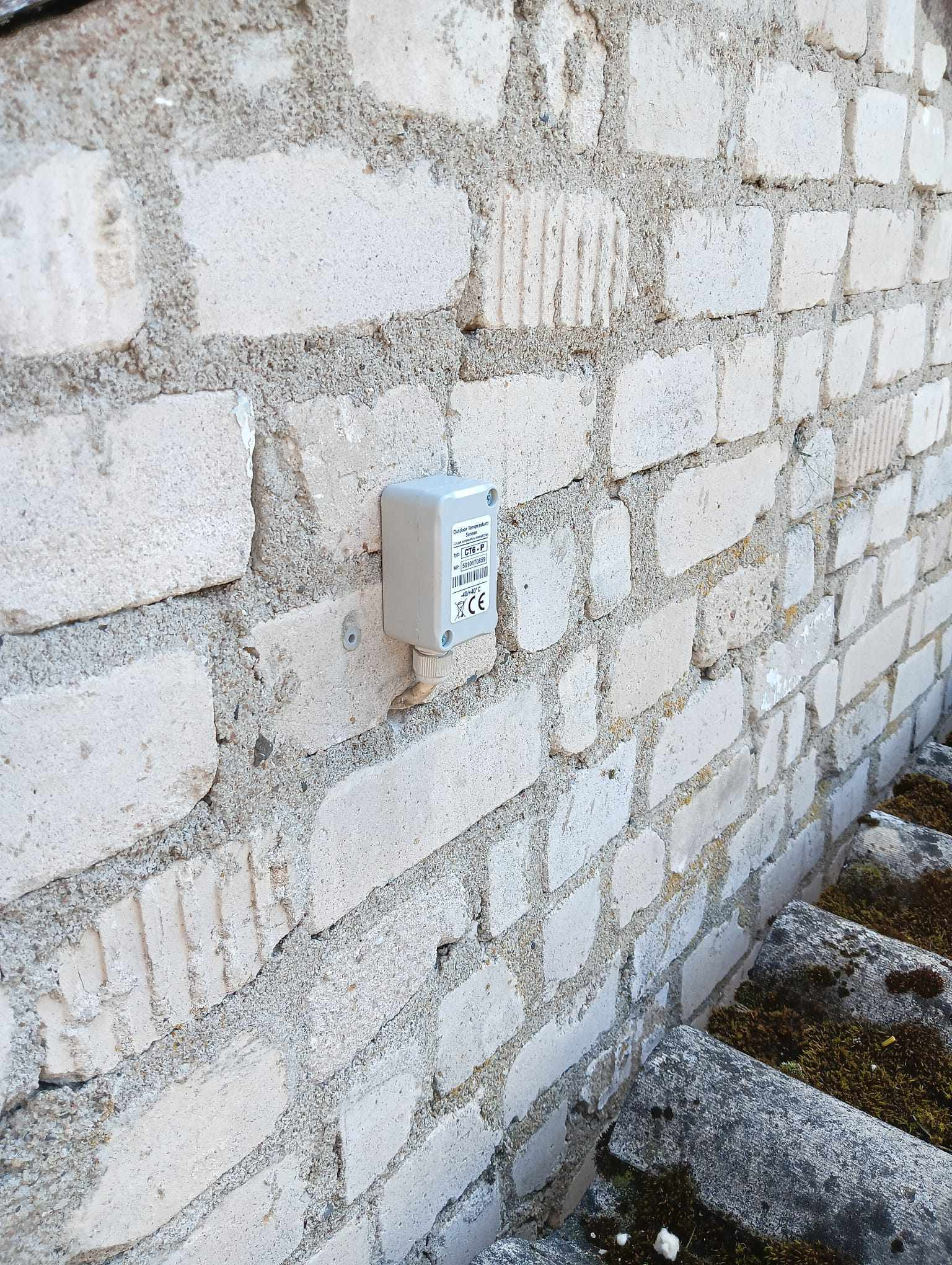 Szara puszka z czujnikiem temperatury zewnętrznej, zamontowana na jasnej, ceglanej ścianie budynku z wystającą, pokrytą mchem krawędzią dachu.
