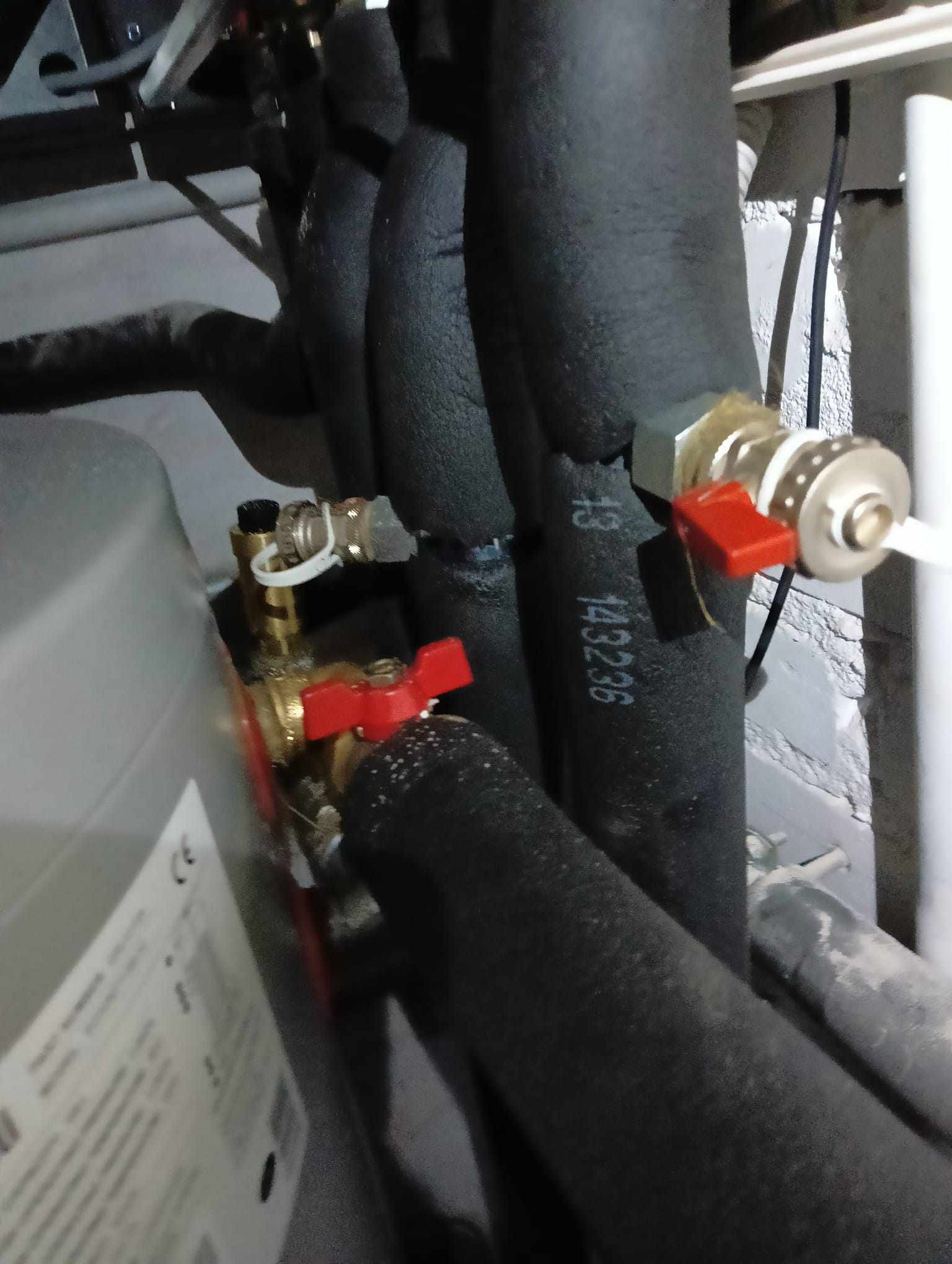 Fragment instalacji z rurami w czarnej izolacji i zaworami z czerwonymi pokrętłami, widoczne numery seryjne na izolacji, w tle szara ściana.