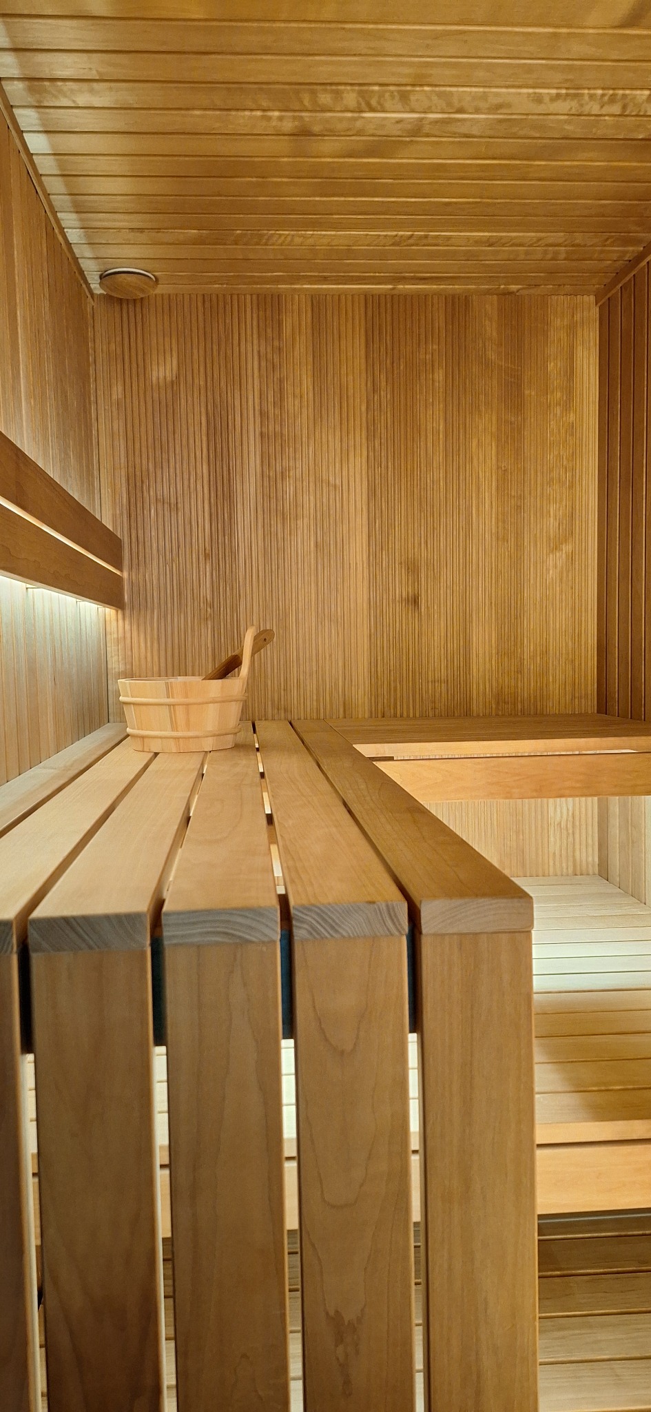 Wnętrze drewnianej kabiny z siedziskami i drewnianym wiadrem z chochlą, oświetlone ciepłym światłem.