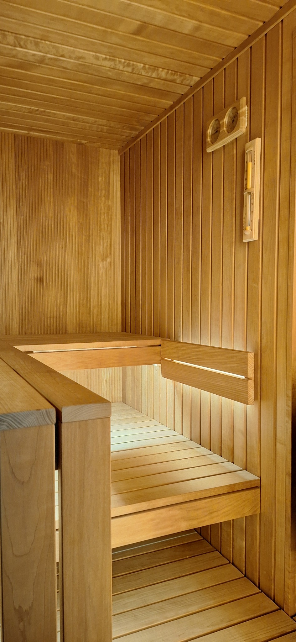 Wnętrze sauny z jasnego drewna z dwupoziomowymi ławkami, oświetleniem LED pod jedną z ławek oraz termometrem i higrometrem na ścianie.