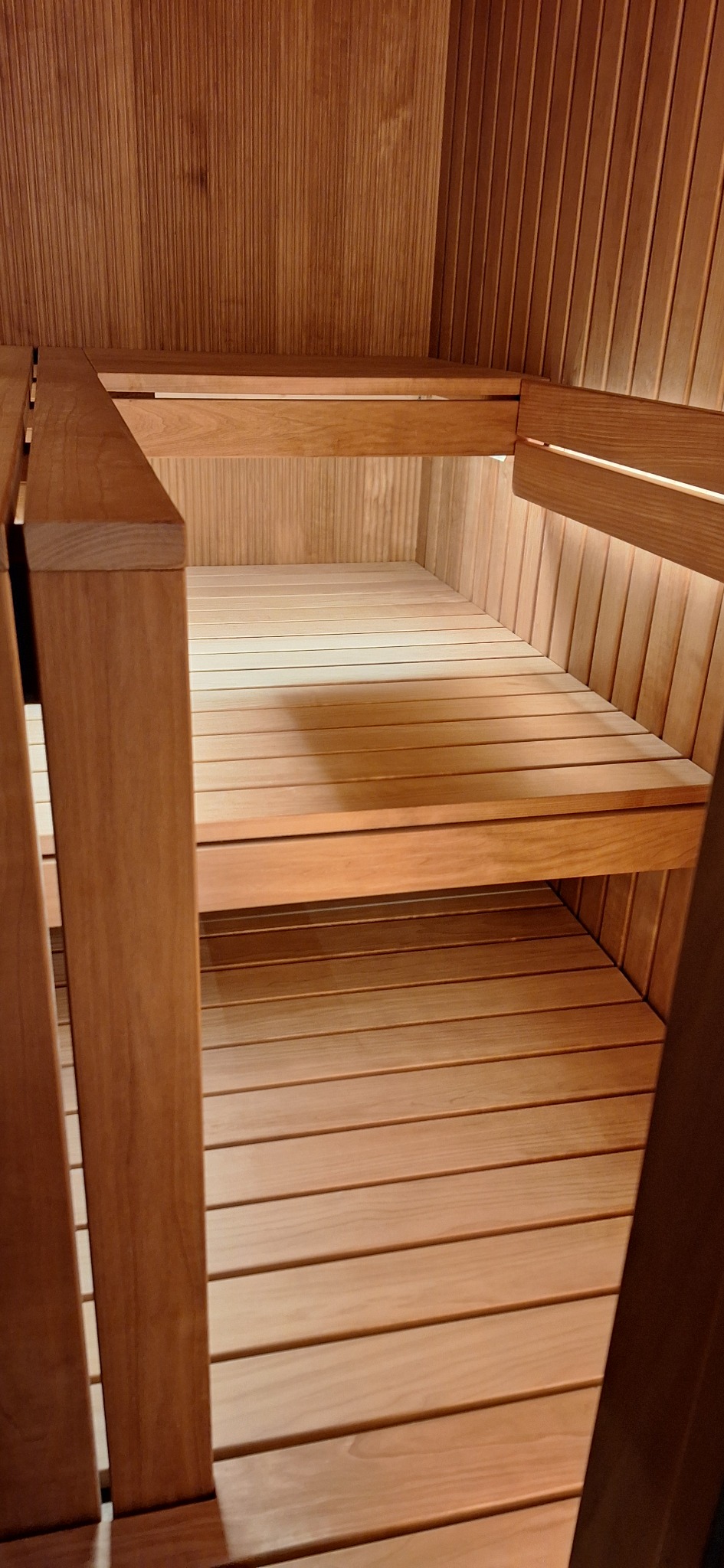 Wnętrze pustej, drewnianej kabiny z dwupoziomowymi ławkami z jasnego drewna, wykonanej z paneli ułożonych pionowo.