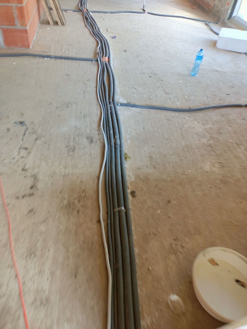 Szereg szarych rur instalacyjnych, częściowo przymocowanych taśmą, ułożonych na surowym, betonowym podłożu w trakcie prac wykończeniowych wnętrza, z widoczną butelką wody i fragmentem muru z cegły.