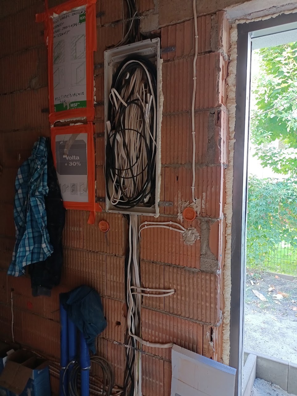 Wewnętrzna instalacja elektryczna w trakcie montażu w surowym budynku z czerwonej cegły, widoczne kable w puszce, przewody prowadzone po ścianie oraz schematy instalacji na ścianie.