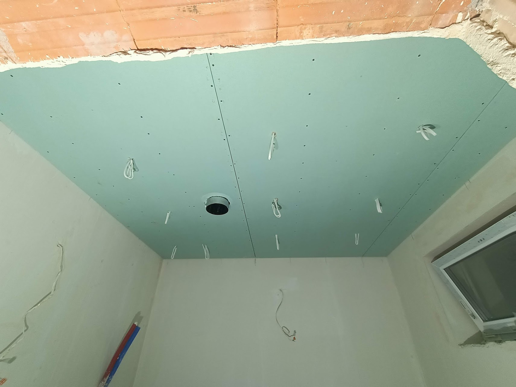 Niebieska płyta kartonowo-gipsowa na suficie z widocznymi przewodami elektrycznymi i otworem pod oświetlenie, w surowym stanie wykończenia wnętrza.