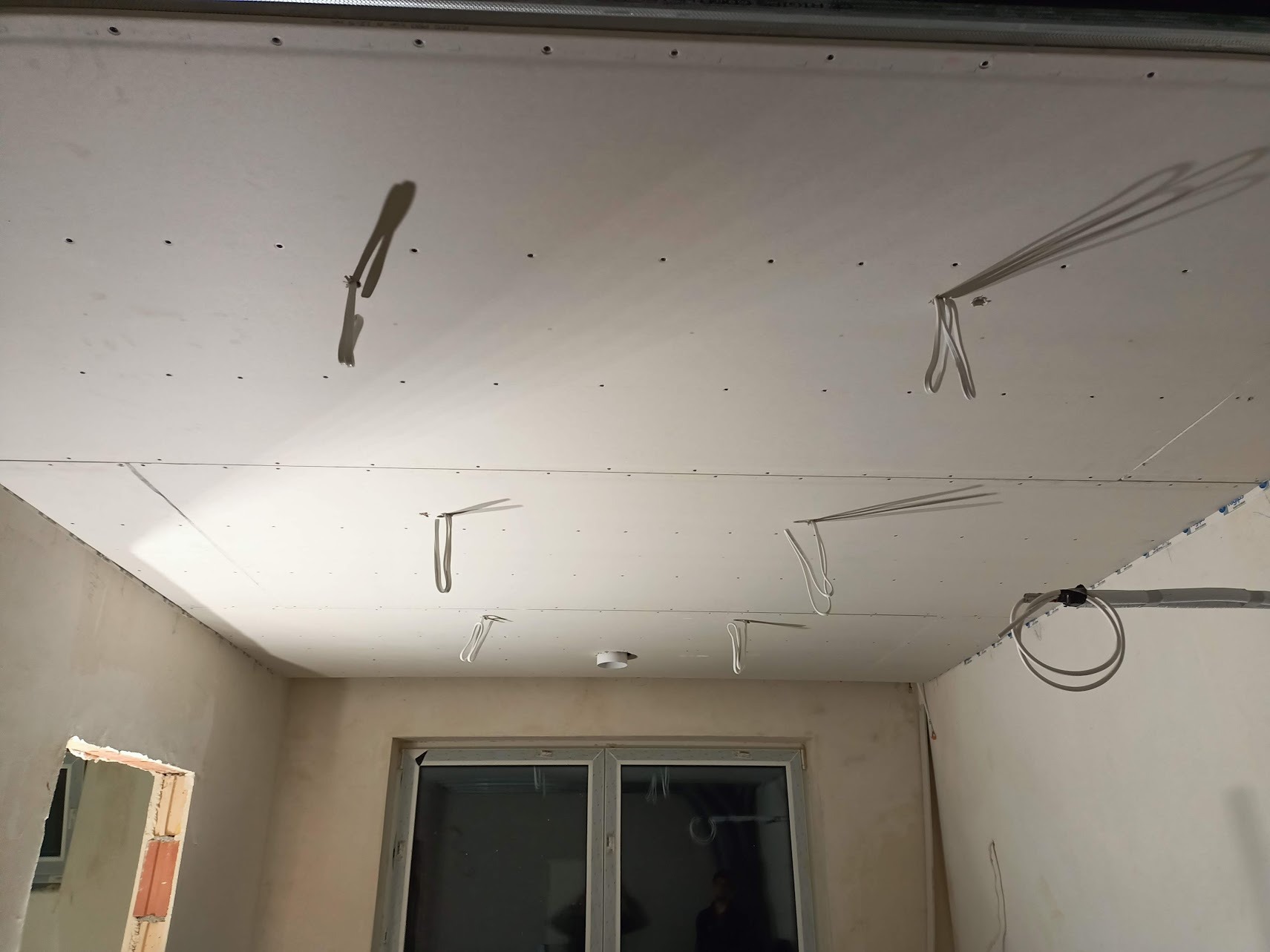 Wnętrze w trakcie prac wykończeniowych: sufit z płyt kartonowo-gipsowych z wystającymi przewodami elektrycznymi, ściany częściowo otynkowane, okno balkonowe.