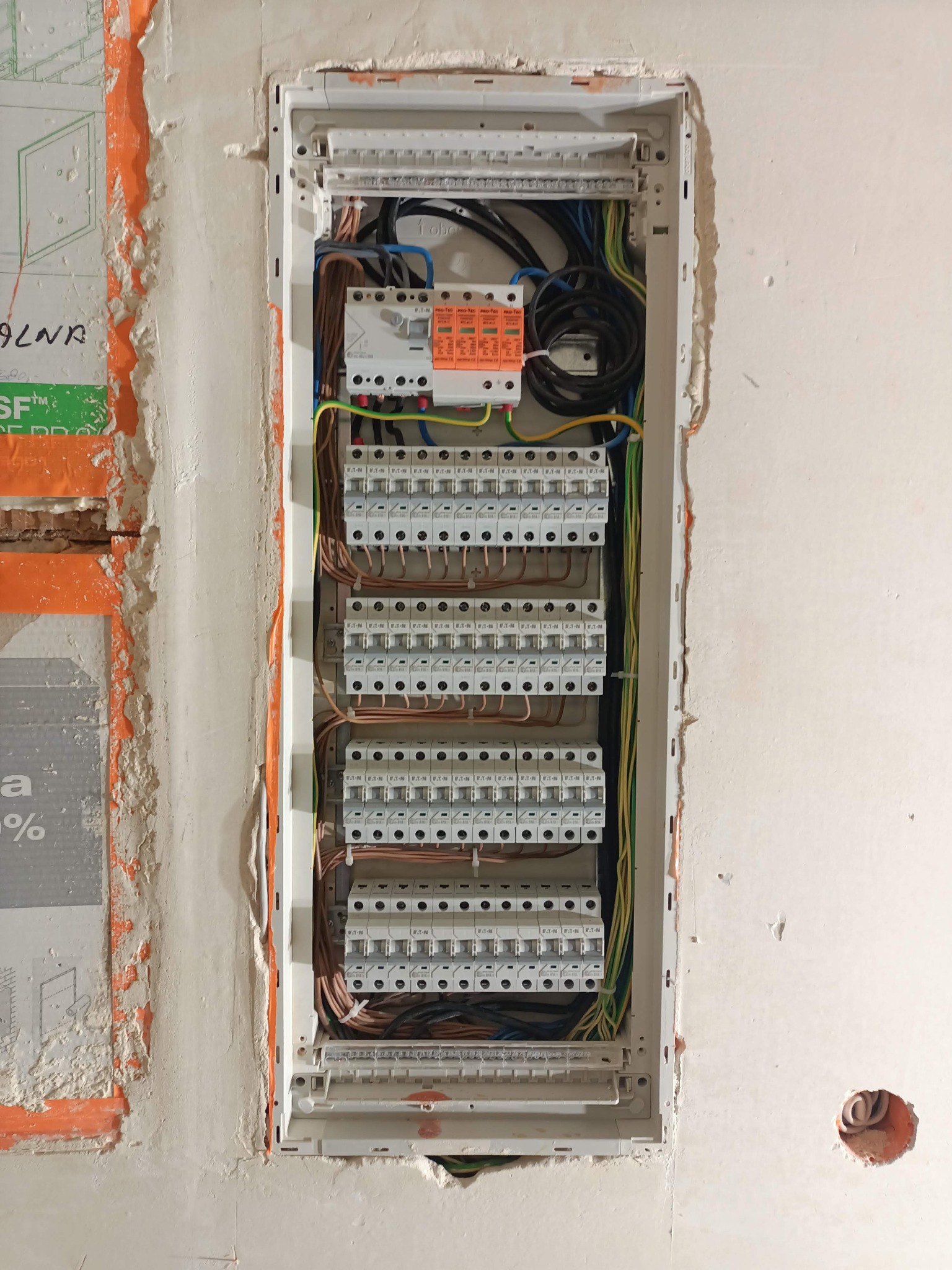 Rozdzielnica elektryczna zamontowana we wnęce w ścianie podczas prac wykończeniowych, widoczne przewody, zabezpieczenia oraz fragment ściany z niedokończonymi pracami gipsowymi.