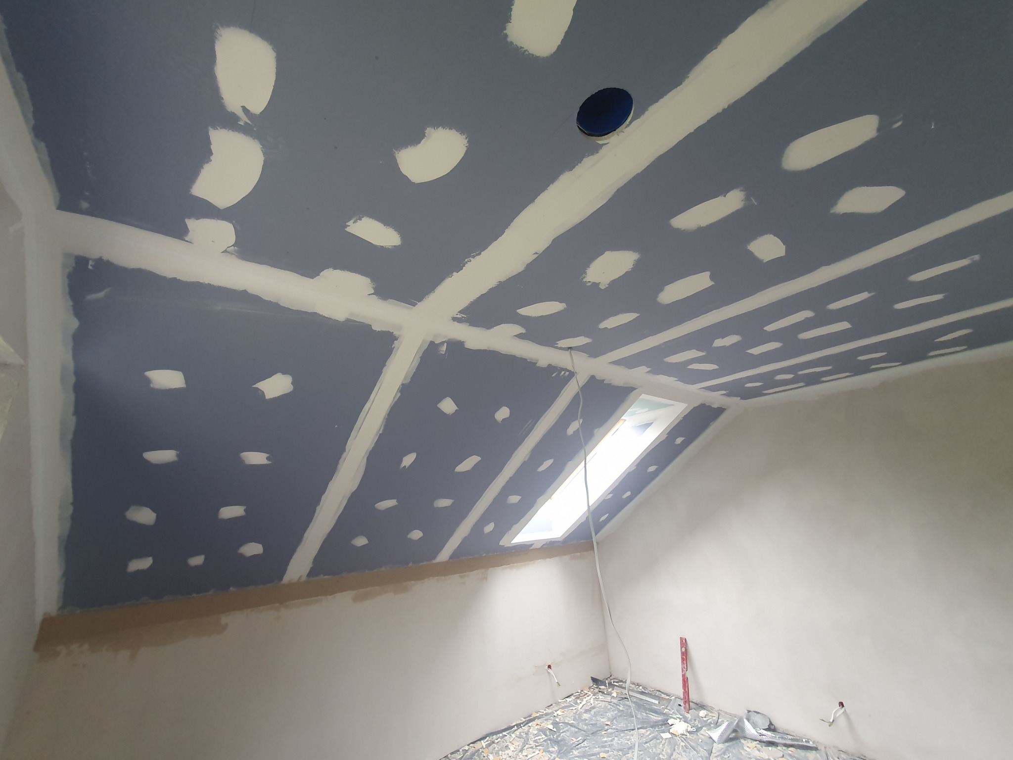 Poddasze w trakcie prac wykończeniowych: szpachlowana płyta gipsowa na skośnym suficie z oknem dachowym i otworem na oświetlenie, widoczne łączenia płyt wzmocnione taśmą.