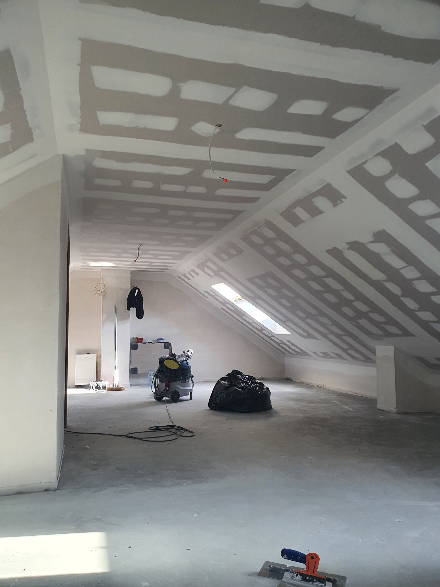 Poddasze w trakcie wykańczania płytami gipsowo-kartonowymi, widoczne szpachlowane łączenia, okno dachowe, narzędzia szpachlarskie na podłodze i odkurzacz przemysłowy.