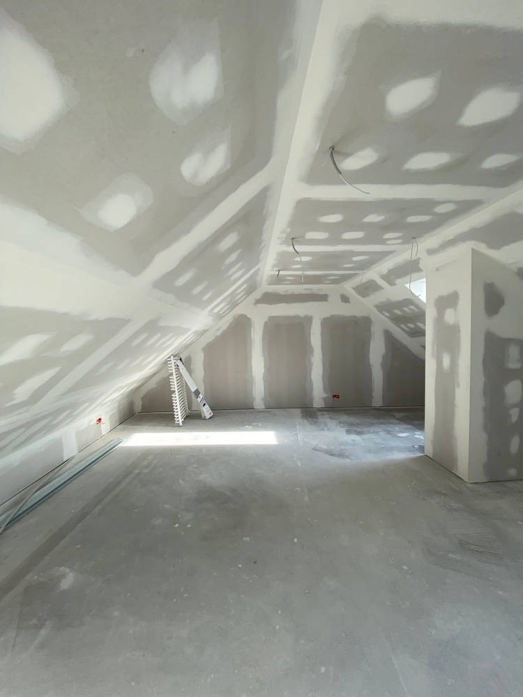 Poddasze w trakcie prac wykończeniowych: widoczne płyty gipsowo-kartonowe na ścianach i suficie, częściowo zaszpachlowane, z widocznymi łączeniami. Na podłodze leży drabina. Wpadające światło...