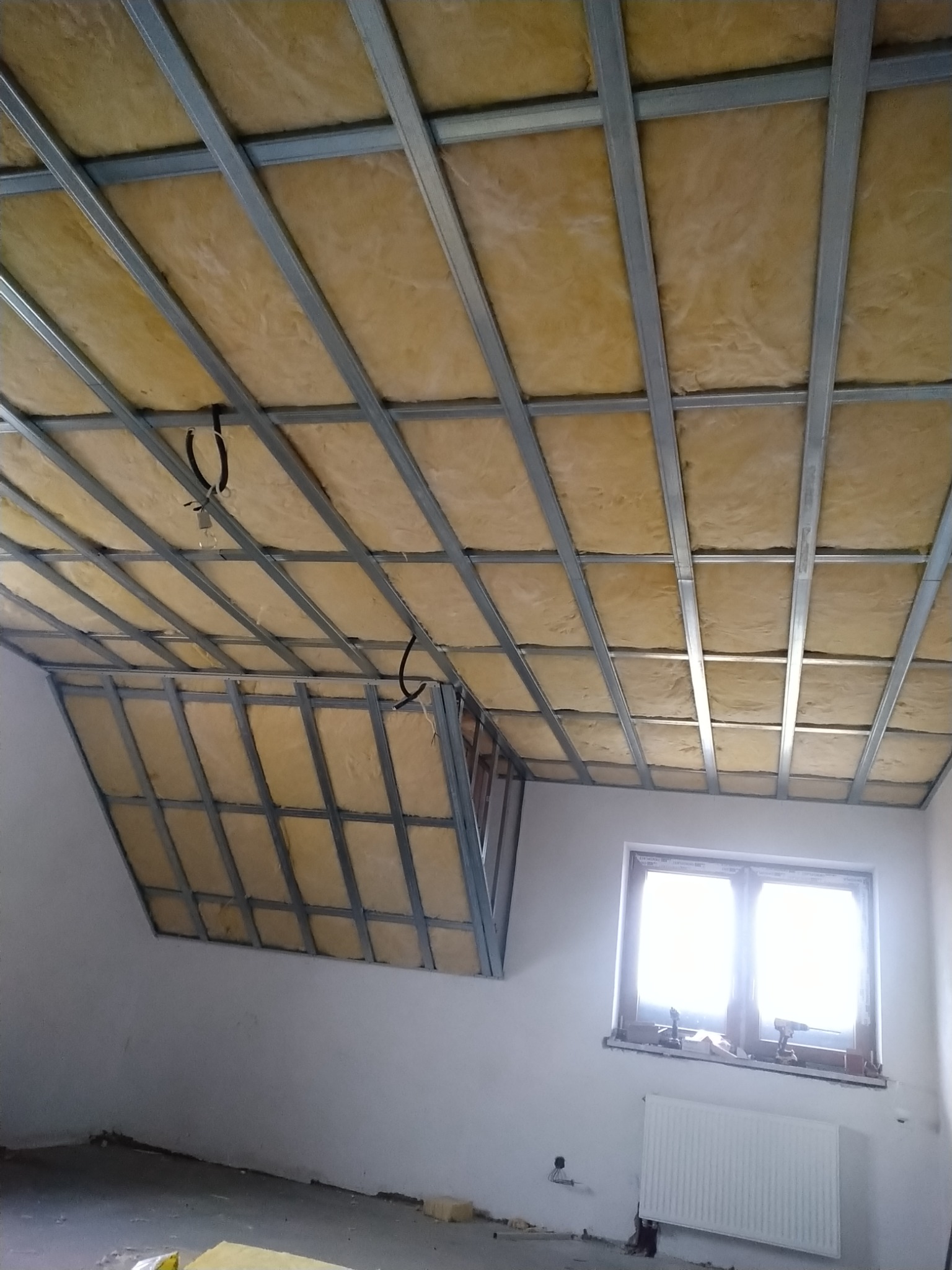 Skośne poddasze w trakcie izolacji wełną mineralną, widoczna metalowa konstrukcja nośna, przewody elektryczne wystające z sufitu, okno z parapetem, grzejnik pod oknem, surowy stan wykończenia.