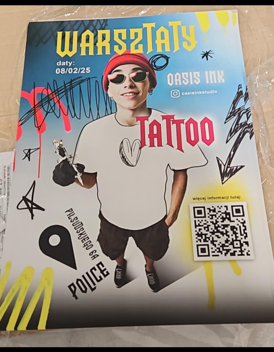 Plakat reklamowy warsztatów tatuażu z wizerunkiem młodego mężczyzny w czapce i okularach, trzymającego maszynkę do tatuażu, z elementami graficznymi i kodem QR.