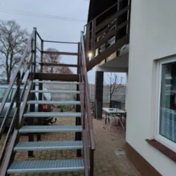 Zewnętrzne, metalowe schody prowadzące na balkon, z kratowanymi stopniami i brązowymi poręczami, na tle domu z oknem i samochodu.