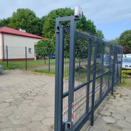 Bartłomiej Lorenc - Nowoczesna, przesuwna brama wjazdowa z szarej siatki panelowej z automatyką, widoczna na tle budynku z czerwonym dachem i zielenią.