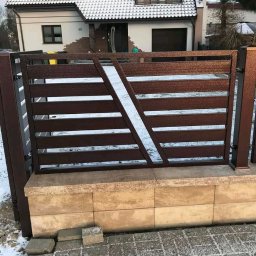 Bartłomiej Lorenc - Nowoczesne, poziome ogrodzenie metalowe w kolorze brązowym z diagonalnym akcentem, zamontowane na niskim murku z jasnego kamienia, na tle ośnieżonego chodnika i domu.