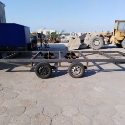 Bartłomiej Lorenc - Metalowa rama przyczepy na dwóch osiach z różnymi felgami, w tle budynek, maszyna budowlana i hałda kruszywa.