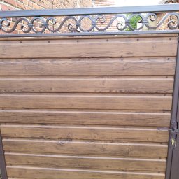 Bartłomiej Lorenc - Brązowa brama z panelami imitującymi drewno i ozdobnym metalowym wykończeniem u góry, widoczny zamek i klamka.