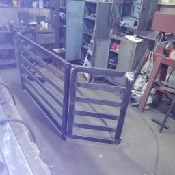 Bartłomiej Lorenc - Surowa, spawana konstrukcja metalowa, przypominająca fragment ogrodzenia lub bramki, widoczna w warsztacie z narzędziami i szafkami w tle.