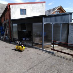Bartłomiej Lorenc - Żółty kompresor Stanley przed częściowo zadaszoną, przeszkloną konstrukcją na posesji z budynkami w tle.